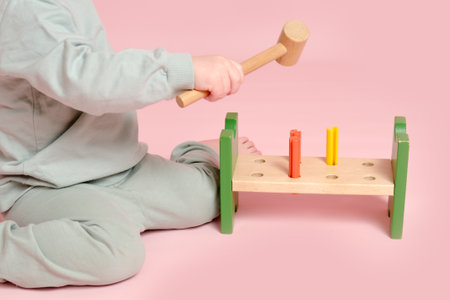 Happy toddler baby plays with a hammer and wooden nails on a studio pink background. Child boy with educational toy. Kid age one year eight monthsの写真素材
