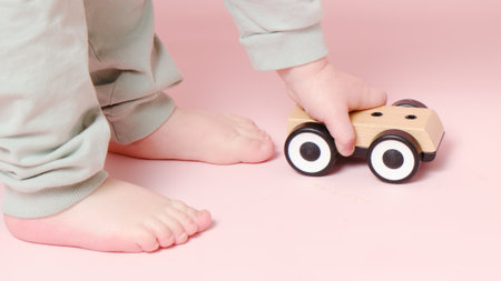 Happy toddler baby is playing with a toy car against a pink background. Child boy rolls a wooden toy car. Kid age one year eight monthsの写真素材