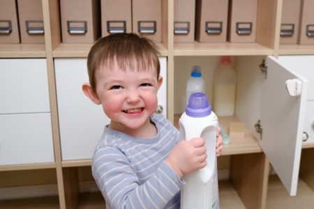 Toddler baby plays with household chemicals and detergent from the closet. Child boy with detergent bottle in home living room. Kid age one year nine monthsの写真素材
