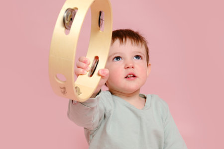 Happy toddler baby with a musical instrument tambourine on a studio pink background. Child boy learning music with percussion instrument. Kid age one year eight monthsの写真素材