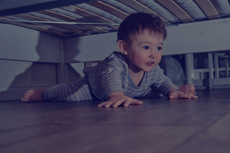 Toddler baby crawls and hides under the bed. Child climbed under the sofa and sits on the floor. Kid boy age one year eight months, full lengthの写真素材