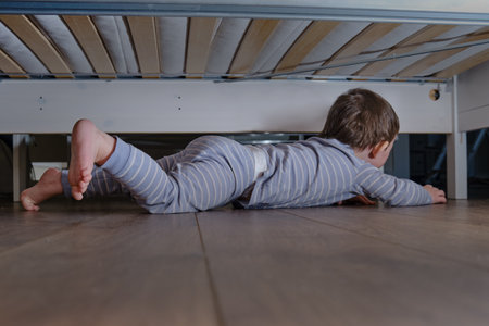 Toddler baby crawls and hides under the bed. Child climbed under the sofa and sits on the floor. Kid boy age one year eight months, full lengthの写真素材