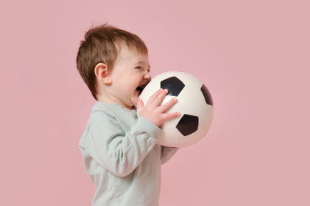Happy toddler baby with a soccer ball on a studio pink background. Child boy holding a sports ball in his hands. Kid age one year eight monthsの写真素材