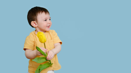 Happy toddler baby with tulip flower on studio blue background, copy space. Smiling child boy with yellow tulip. Kid age one year eight monthsの写真素材