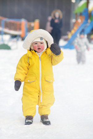 Portrait of a toddler baby in warm yellow clothes on a winter playground. Happy baby boy in snowsuit standing in the snow in kindergarten. Kid age one year eight monthsの写真素材