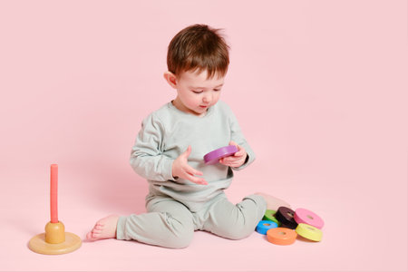 Happy toddler baby plays with a pyramid on a studio pink background. Child boy puts wooden rings on a toy pyramid. Kid age one year eight months, full heightの写真素材