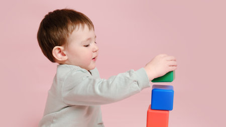Happy toddler baby plays with cubes on studio pink background. Child boy builds a tower of toys. Kid age one year eight monthsの写真素材