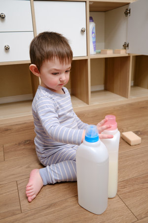 Toddler baby plays with household chemicals and detergent from the closet. Child boy with detergent bottle in home living room. Kid age one year nine monthsの写真素材