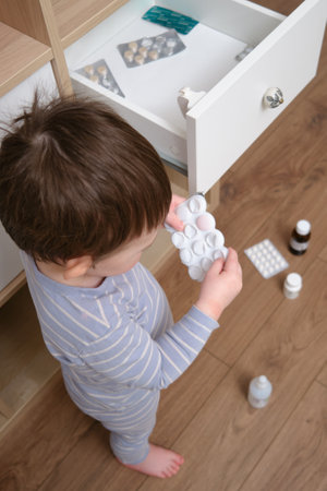 Toddler baby opened the cabinet drawer with pills and medicine. Child boy holding a pack of pills in the home living room. Kid age one year nine monthsの写真素材