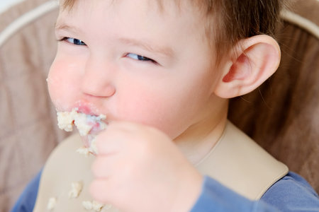 A happy child eats porridge with a spoon while sitting on a high chair. Baby in a bib eats oatmeal porridge himself. Kid aged about two years (age one year nine months)の写真素材