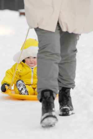Toddler baby rides in the snow on icesled, a winter playground. Mother woman sledding baby boy in yellow snowsuit. Kid age one year eight monthsの写真素材