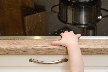Toddler baby climbs on a hot electric stove in the home kitchen. A small child touches the surface of the stove with his hand at the risk of getting burned. Kid aged one year eight monthsの写真素材