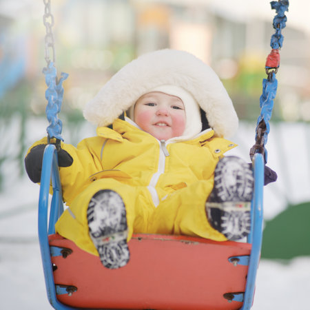 Happy toddler baby rides on a swing on a winter playground. Baby boy in yellow snowsuit swinging in kindergarten. Kid age one year eight monthsの写真素材
