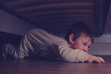 Toddler baby crawls and hides under the bed. Child climbed under the sofa and sits on the floor. Kid boy age one year eight months, full lengthの写真素材