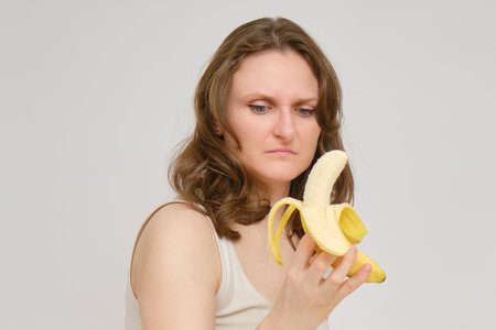 A woman with doubt and contempt looks at a banana, a studio white background. A disgusted look at the banana fruit in the hands of a woman.の写真素材