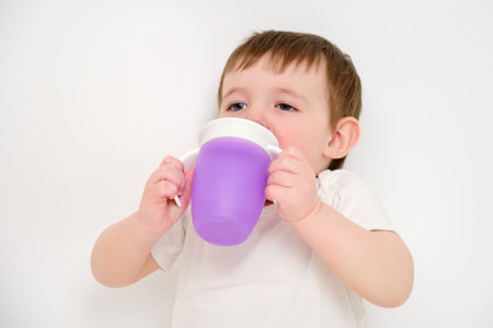 Happy baby drinks water from cup on studio white background. Resting child with cup juice in hands. Kid about two years old (one year nine months)の写真素材