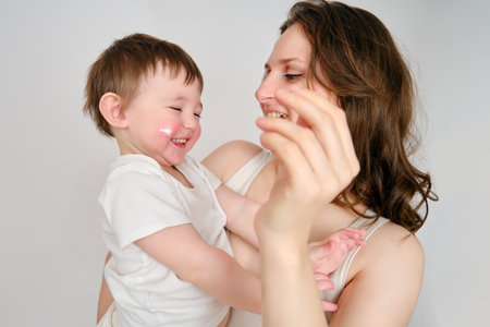 Woman mother smears her cheeks with baby cream on a studio white background. Mom smears child's face with a moisturizer for eczema. Kid about two years old (one year nine months)の写真素材