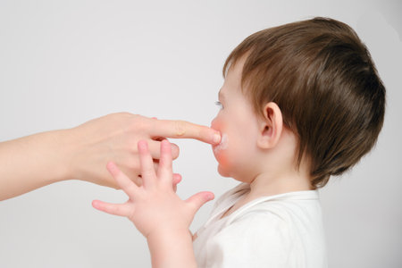 Woman mother smears her cheeks with baby cream on a studio white background. Mom smears child's face with a moisturizer for eczema. Kid about two years old (one year nine months)の写真素材
