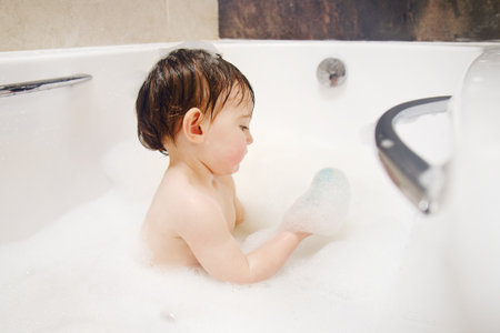 A happy child is playing with toys while sitting in a white homemade bathtub. Little toddler baby boy bathes in a bubble bath. Kid aged one year six monthsの写真素材