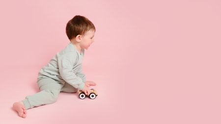 Happy toddler baby is playing with a toy car against a pink background. Child boy rolls a wooden toy car. Kid age one year eight months, full heightの写真素材
