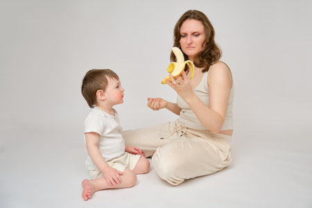 Happy baby with mother eating banana on studio white background. Portrait of a smiling child with mom snacking on fruits. Kid about two years old (one year nine months)の写真素材