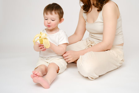 Happy baby with mother eating banana on studio white background. Portrait of a smiling child with mom snacking on fruits. Kid about two years old (one year nine months)の写真素材