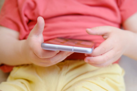 The little boy uses his finger to touch the screen of the mobile device while having fun playing a game on the brown couch. Playing games on the smartphone is a popular pastime for childrenの写真素材