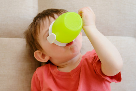 The little baby happily holds a mug filled with water, sipping on the white liquid while lounging on the sofa. Drinking from a cup, the child smiles on the couch. Kid aged about two yearsの写真素材