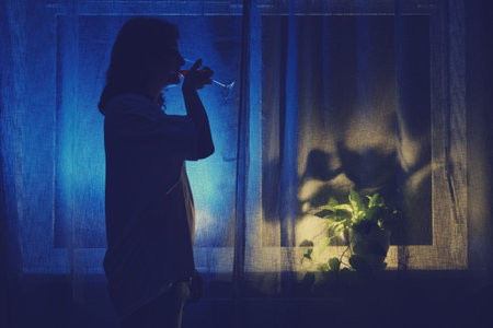 A woman drinks wine from a glass at the night dark window, low key silhouette. Problems of female...