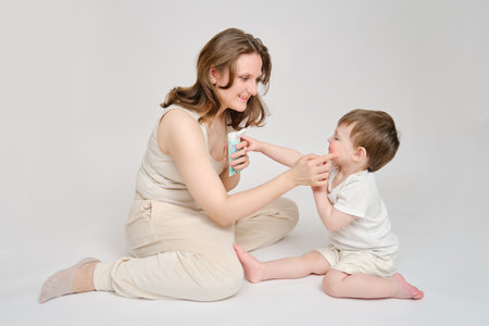 Woman mother smears her cheeks with baby cream on a studio white background. Mom smears child's face with a moisturizer for eczema. Kid about two years old (one year nine months)の写真素材