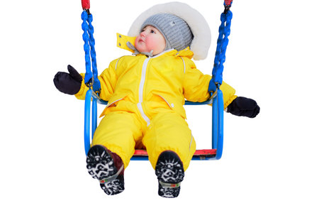 Toddler baby boy in a yellow snowsuit is swinging on a swing in a snowy playground, isolated on white background. Child age one year five months playing on a winter walkの写真素材
