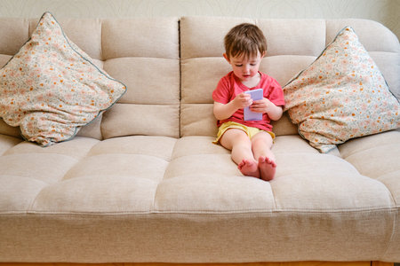 The content child is sitting on the couch with a phone in hand. The delighted kid lounges on the...