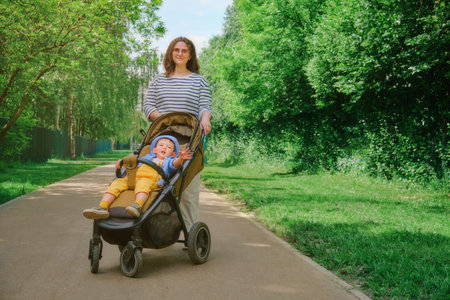 The child and mother are taking a leisurely stroll through the park with their carriage. Kid aged...