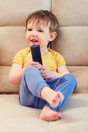 The joyful child is seated on the sofa holding the TV remote, watching cartoons with a beaming smile. A little baby is lying on the couch, watching a cartoon on the television set with happiness.の写真素材