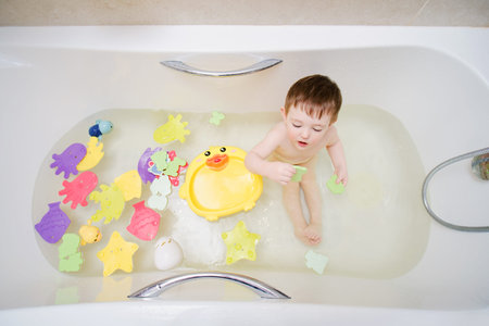 A happy baby is washing in a white bathtub with bathing toys. Funny child in the home bathroom. Kid...