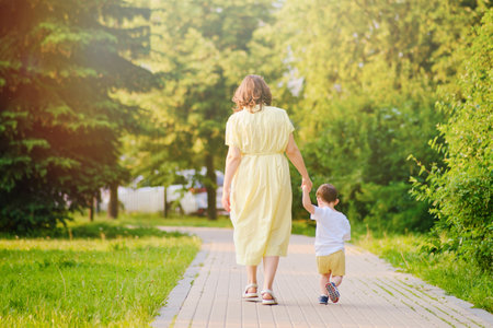 A child takes steps holding a mother woman by the hand along a path in nature. Happy baby with mom on a walk in the summer park. Kid aged about two years (one year eleven months)の写真素材