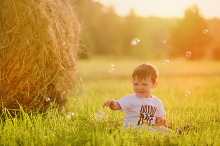 A happy child, a toddler, enjoys playing with soap bubbles on the field, while the straw stacks stand in the background. Portrait of a happy kid aged two yearsの写真素材