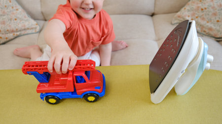 The baby must refrain from playing with a hot iron in the living room due to the potential danger it poses. Children need to be carefully monitored to avoid any incidents. Kid aged about two yearsの写真素材