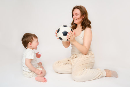 Happy baby with mother play with fotball ball on studio white background. Portrait of a smiling child with mom and playing while sitting on the floor. Kid about two years old (one year nine months)の写真素材