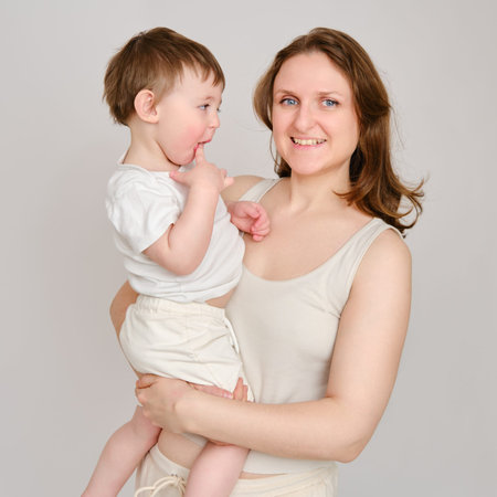 Happy baby in the arms of a mother woman on a studio white background. Portrait of a smiling child with mom. Kid about two years old (one year nine months)の写真素材