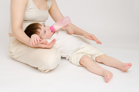 Happy baby with mother drinks milk from bottle on studio white background. Resting child with mom eats formula. Kid about two years old (one year nine months)の写真素材