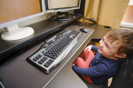 Toddler baby works on a computer with a keyboard and mouse. Happy child sits on a computer chair next to the PC monitors. Kid age one year nine monthsの写真素材