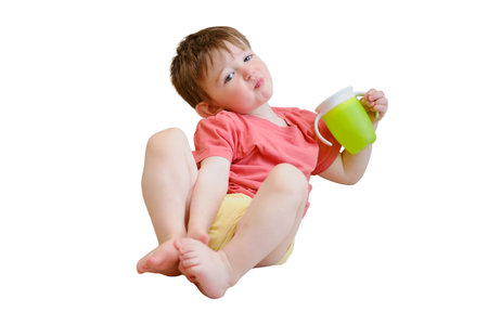 The non-spill cup is in the hands of the smiling child, who sits contentedly on the couch, drinking water, isolated on white background. Kid aged about two yearsの写真素材
