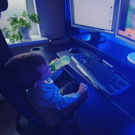 Happy baby is sitting at the computer keyboard with a bottle of milk in his hands. A child with a desktop PC in a home living room drinks milk formula from a bottle. Kid aged one year nine monthsの写真素材
