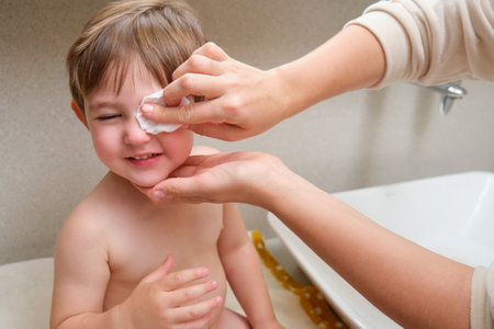 A woman mother cleans her child eyes with a cotton pad in the home bathroom. Kid aged two years (two-year-old boy) Mom washes toddler baby eyesの写真素材