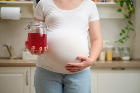 A pregnant woman holds a jar of juice in her hands while standing in her home kitchen. Pregnancy and proper nutrition while expecting a childの写真素材
