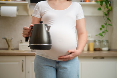 A pregnant woman holds an electric kettle in her hands, home kitchen background. Pregnancy and proper nutrition while expecting a babyの写真素材