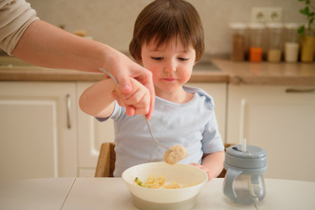 Mother woman teaches toddler baby to eat with a fork in home kitchen. Mom and son are cooking together in the kitchen. Toddler baby two years old at dinnerの写真素材