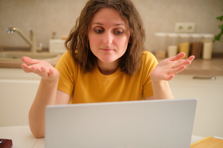 A woman with a laptop spreads her arms while sitting at a table in a home kitchen. An adult female...