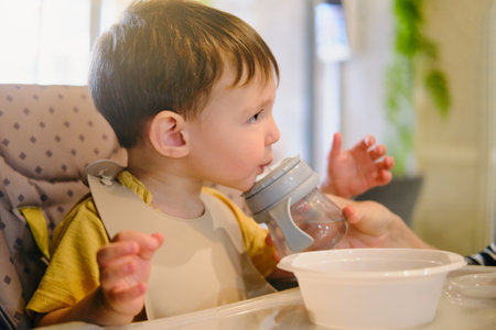 Mother woman helps baby boy drink water from a bottle of non-spill bottle in a cafe. A child eats in a restaurant sitting at a table on a high chair. Kid aged two yearsの写真素材